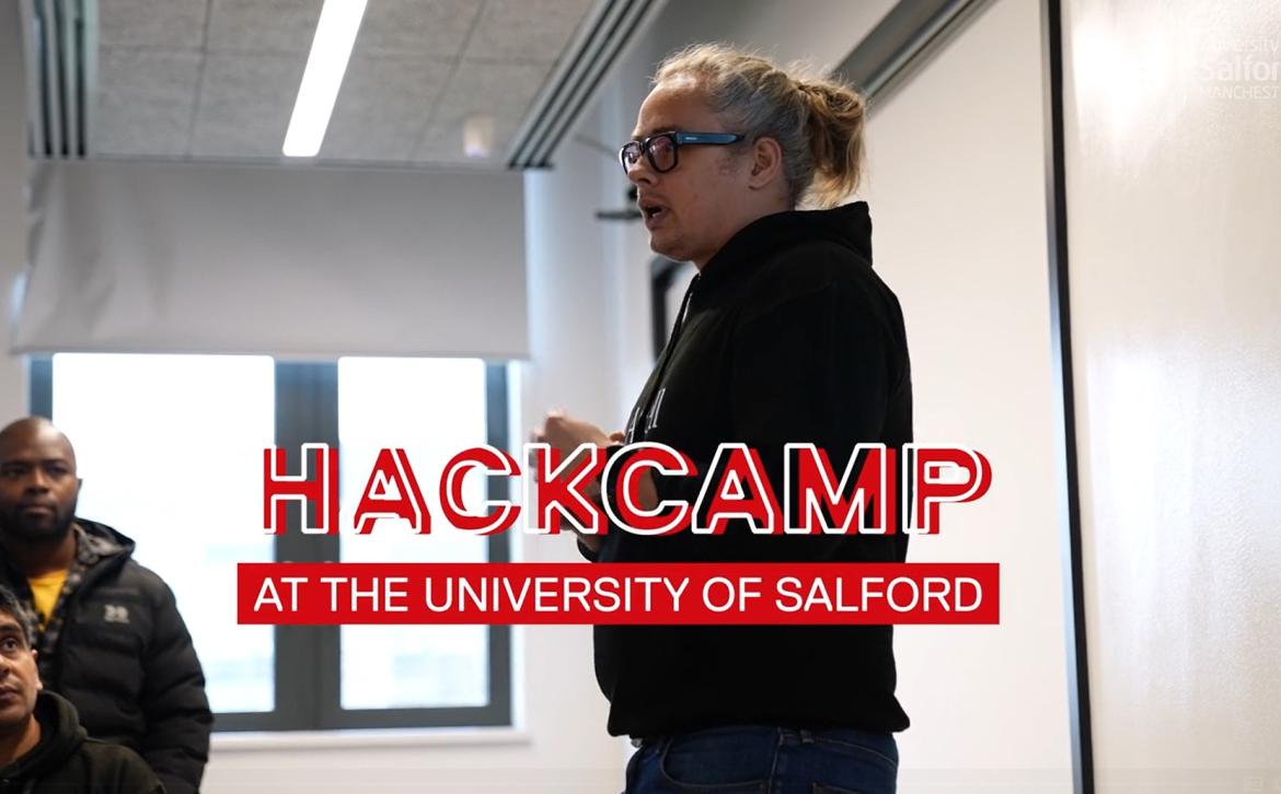 A man wearing an AJ Bell hoody doing a presentation. The words 'Hackcamp at the University of Salford' are overlaid