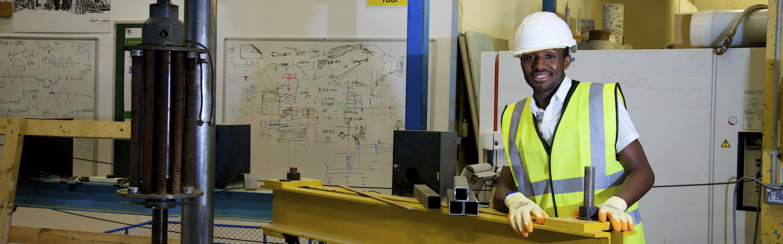 A student stood in our Heavy Structures lab with his hands on a metal beam and smiling while wearing a yellow hi-vis vest and a white hard hat