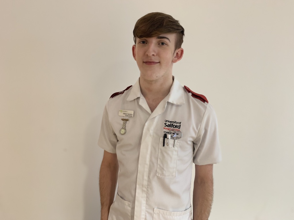 Nathan Harrison is smiling at the camera wearing a white University of Salford nurse uniform
