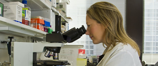 Woman in lab coat looks down a microscope