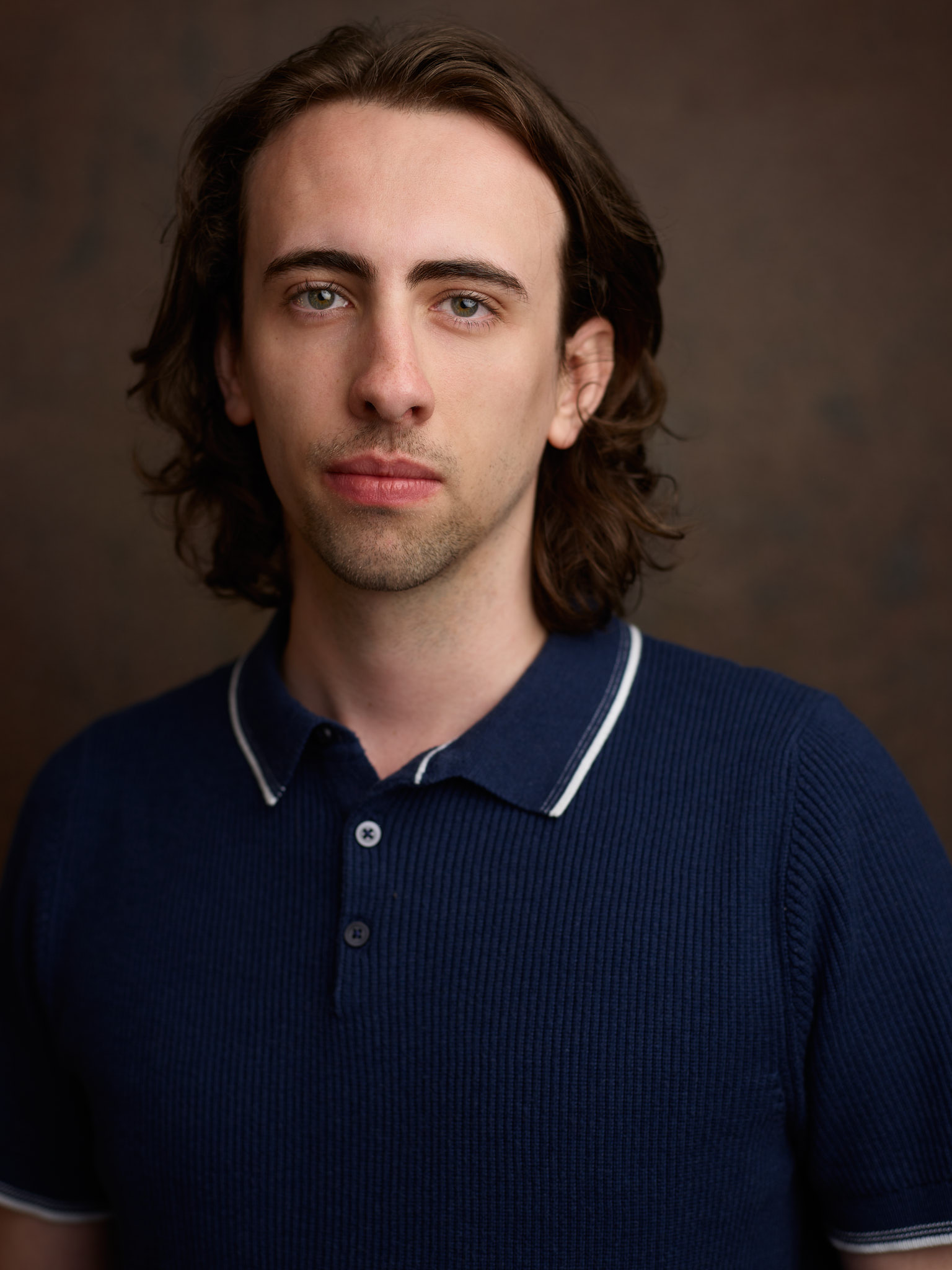 Actors headshot - a man with shoulder length brown hair wears a blue collared tshirt