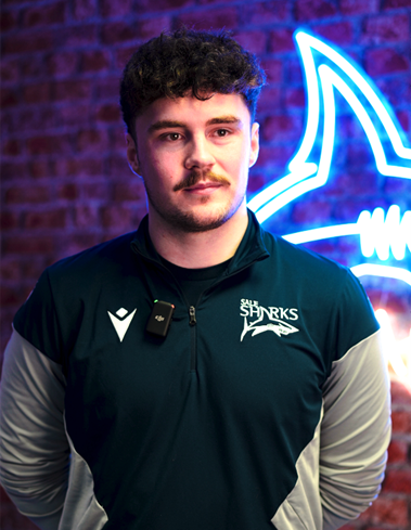 Man in blue and grey 'Sale Sharks' sweatshirt stood in front of a brick wall with a neon shark sign hanging on it. 