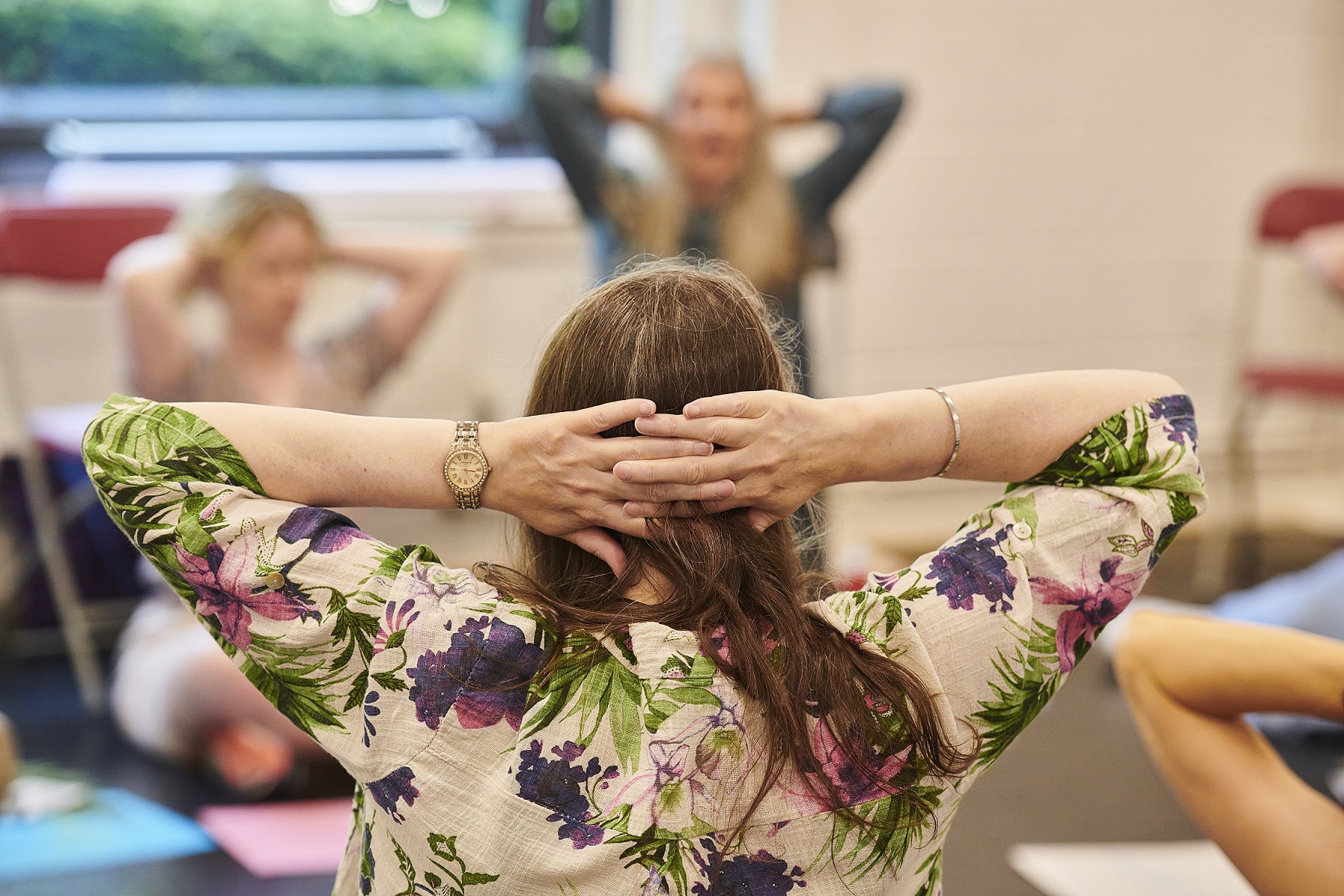 A group of women are sat on the floor in a circle with their hands on their heads, taking part in a workshop.