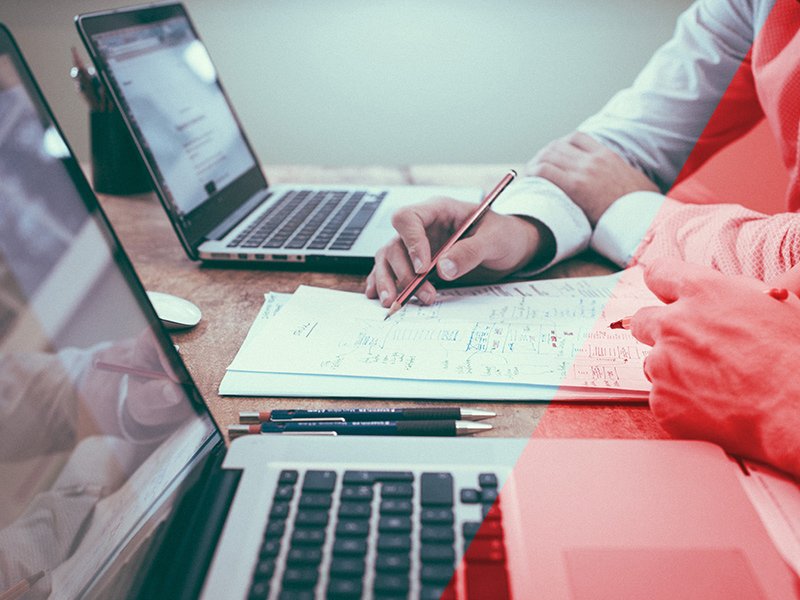 Two people sat with laptops looking at notes on paper together.