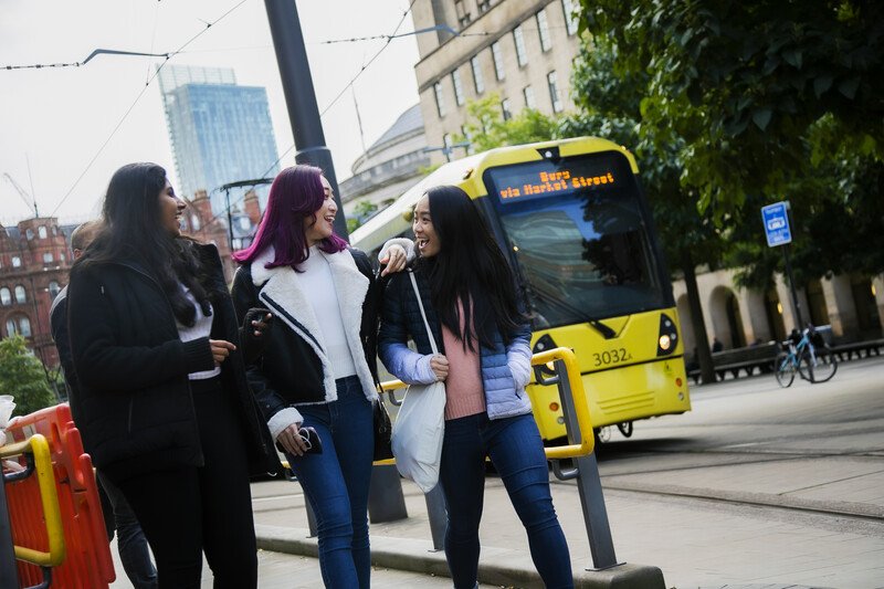 Three students walking past a tram in Manchester City Centre