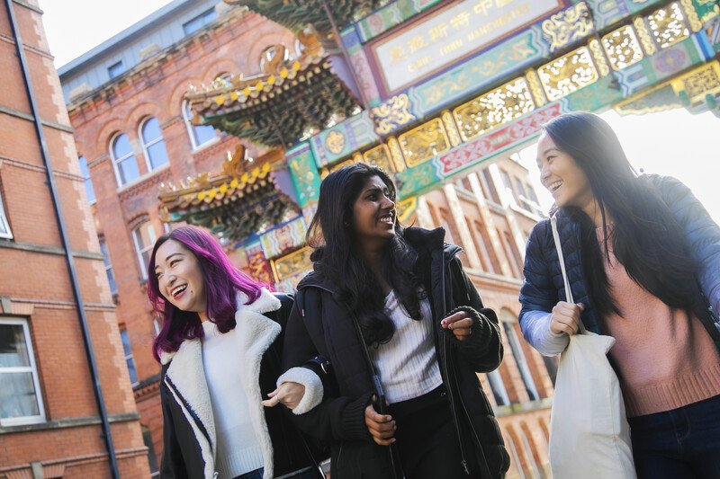 Three female students chatting and laughing while walking through Chinatown, with Chinese archway in the background