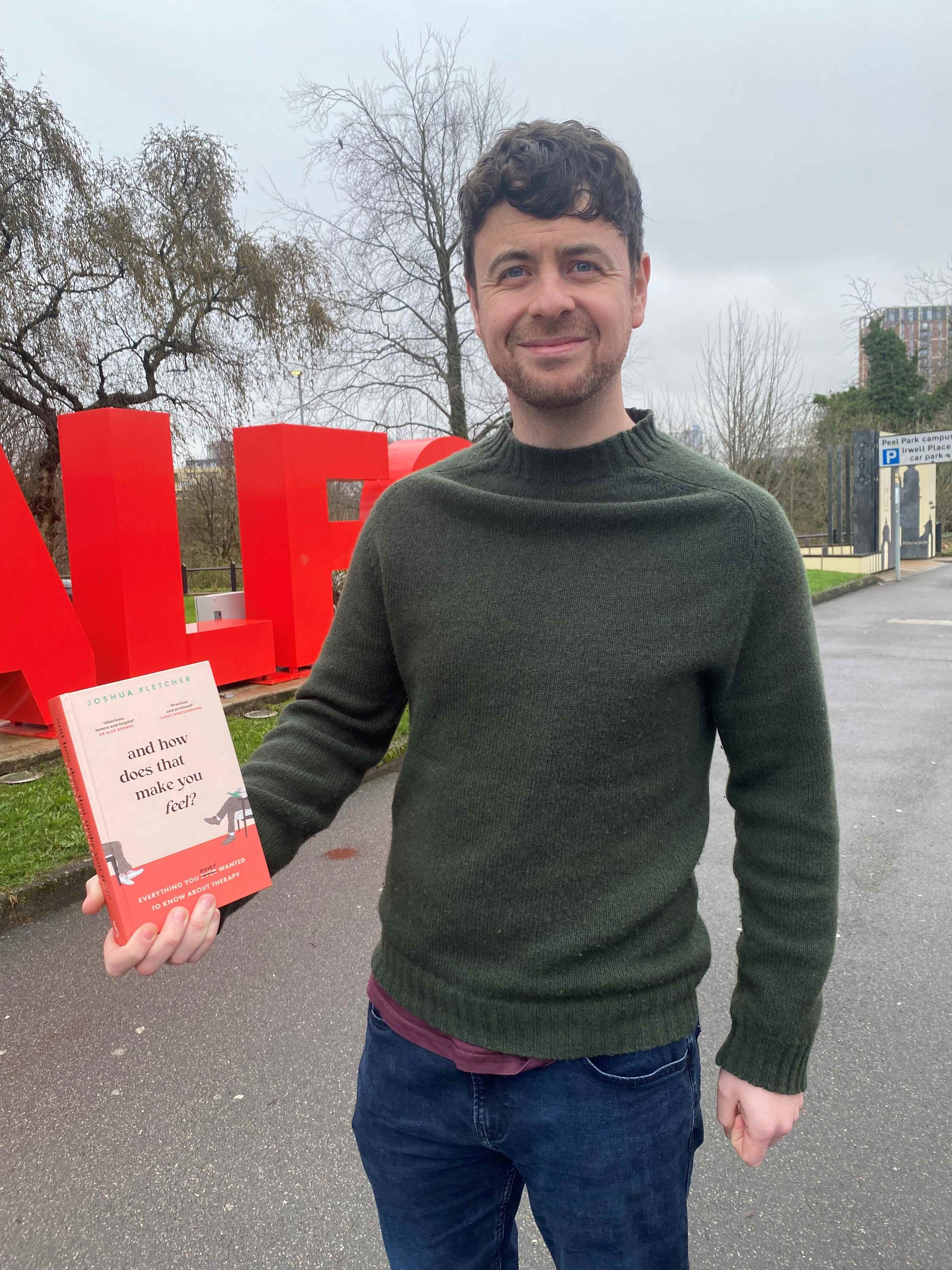 Image shows Josh holding his book in front of the Salford sign