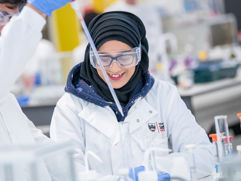 Science student wearing a University of Salford lab coat, watching another student using a pipette in a test tube