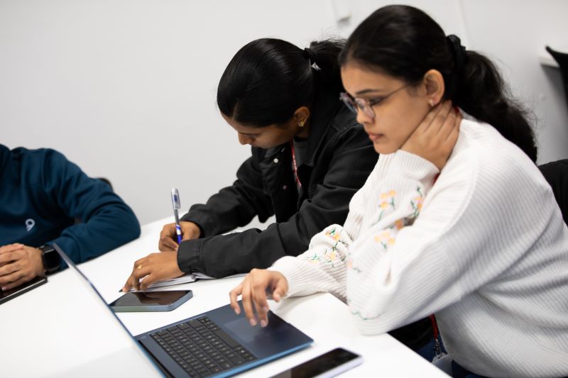 Three seated students with a laptop and mobile on a desk