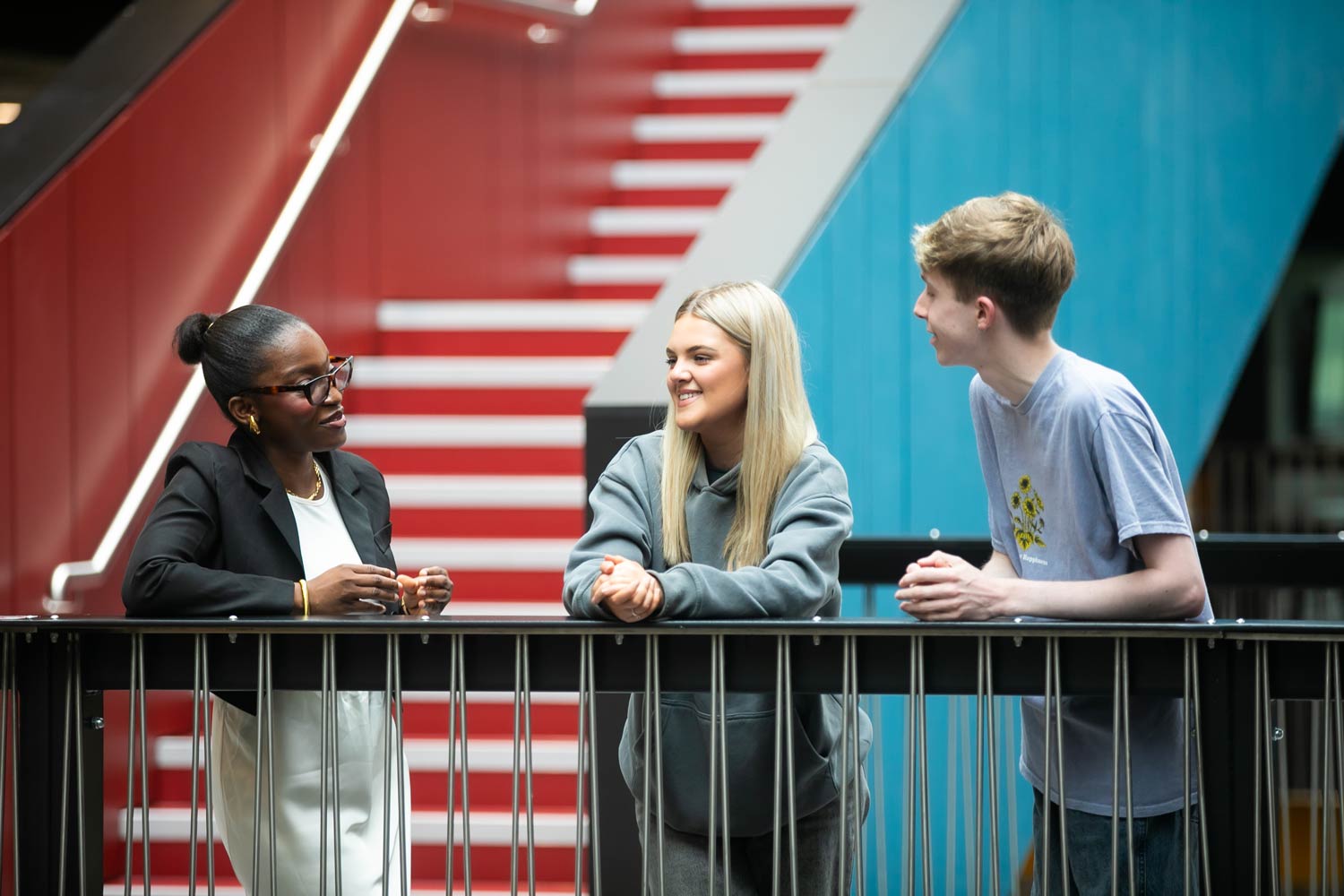 Three students engaged in conversation while leaning on a railing,