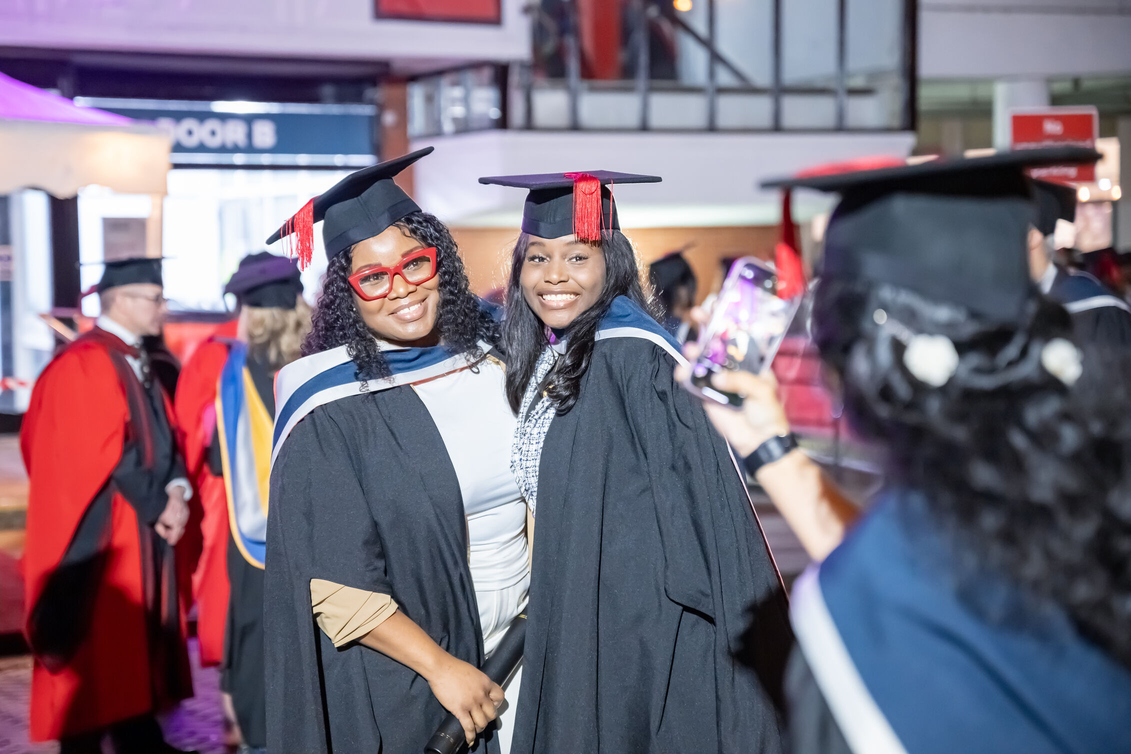 Two friends in graduation robes posing for a photograph outside Maxwell Hall, University of Salford