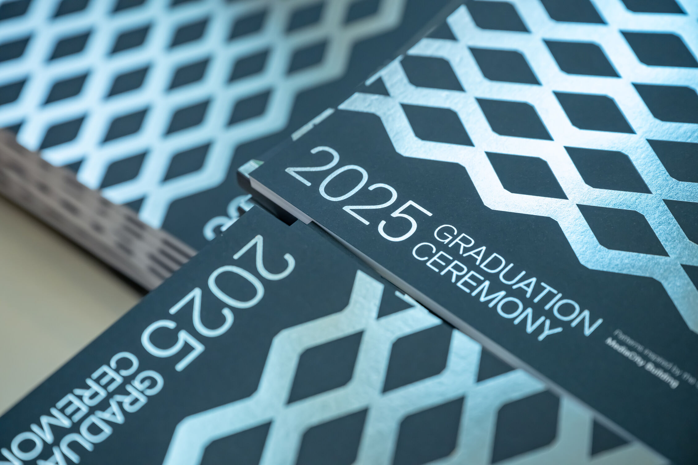 A pile of navy blue graduation brochures. The brochures read 2025 graduation ceremonies in silver foil writing and have a diamond pattern covering them also in silver foil