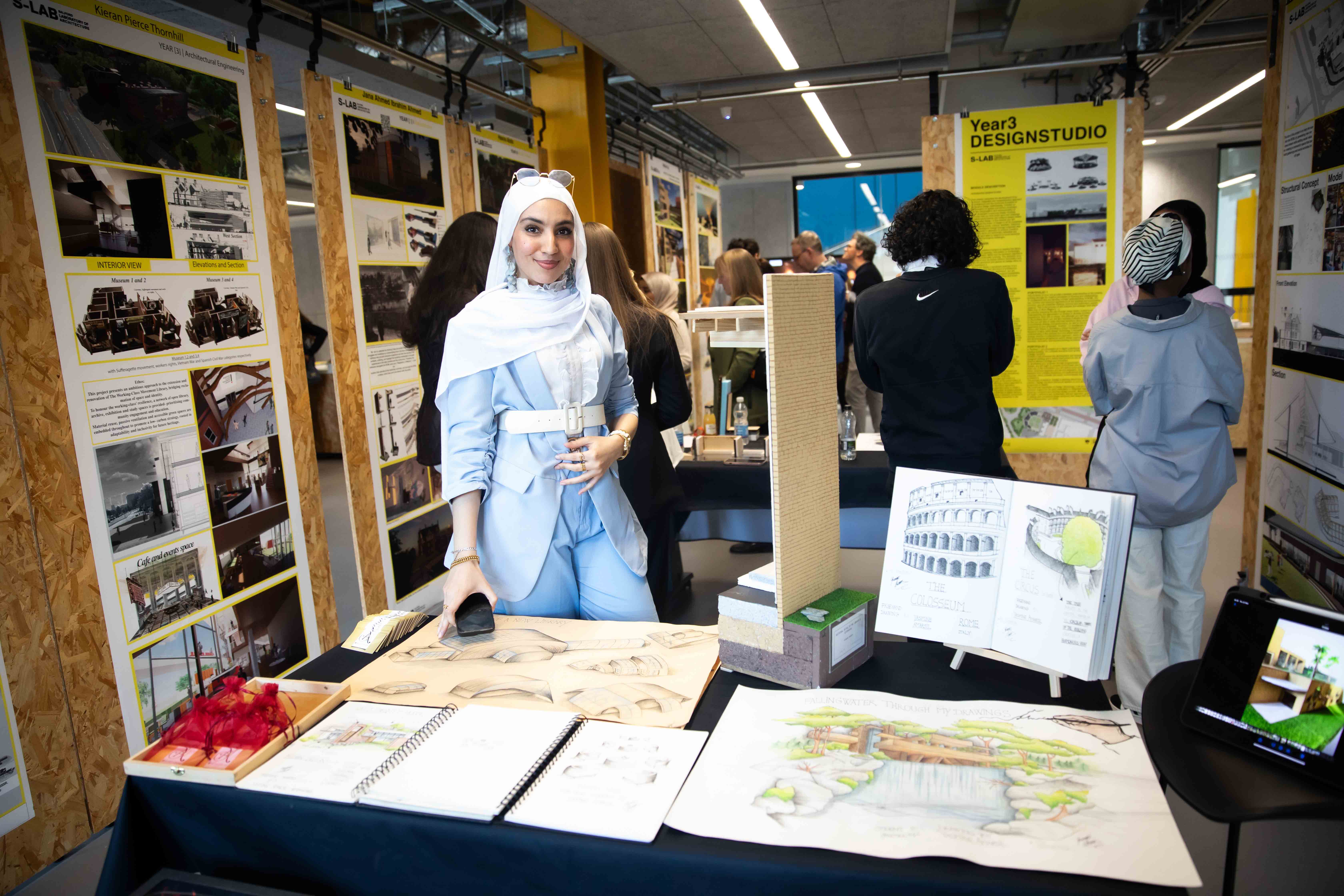 At our Architecture Degree Show, a student stands behind a table displaying her work, which includes a selection of sketches, design plans and scale models.