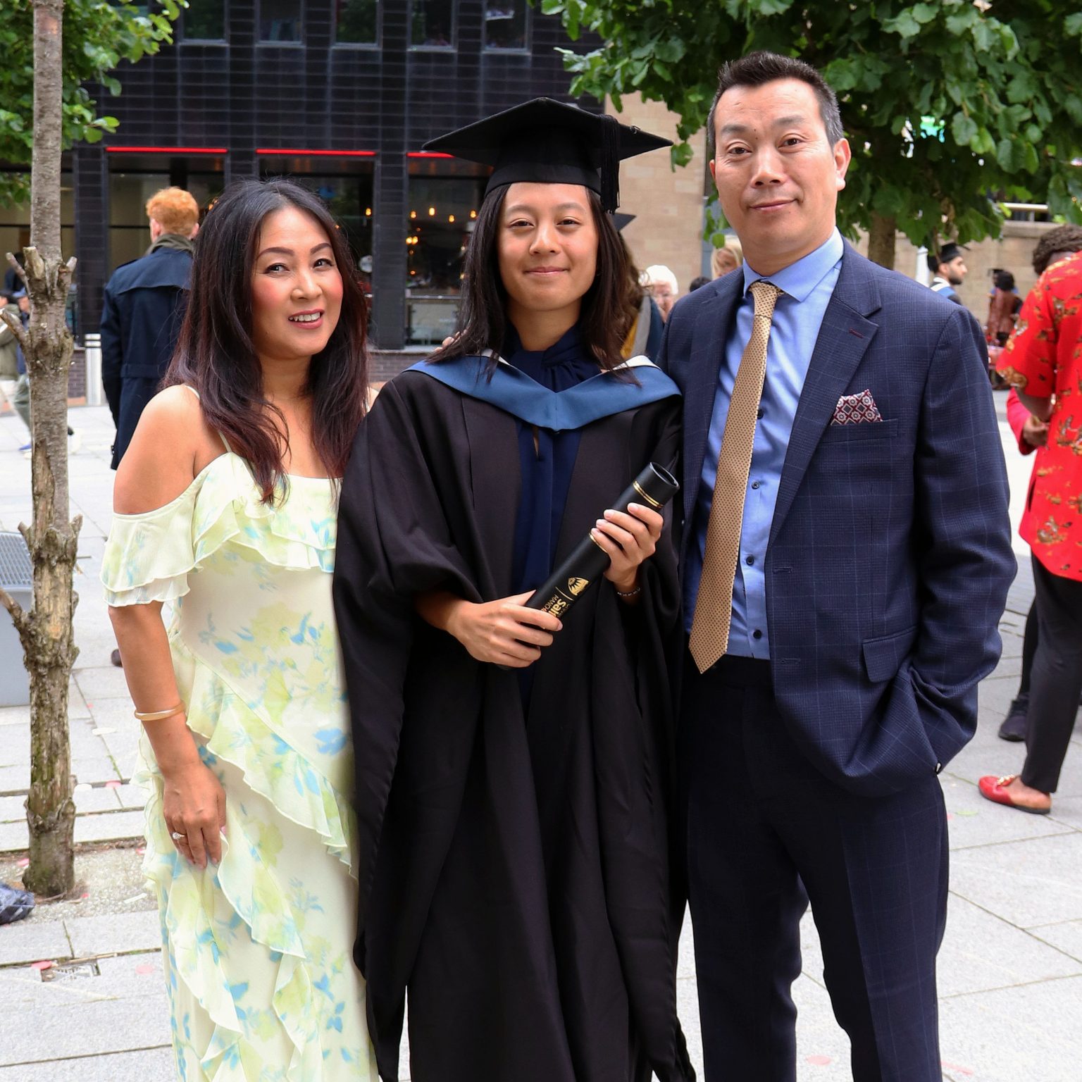 Megan, dressed in a graduation cap and gown, stands outdoors smiling with her parents on her graduation day