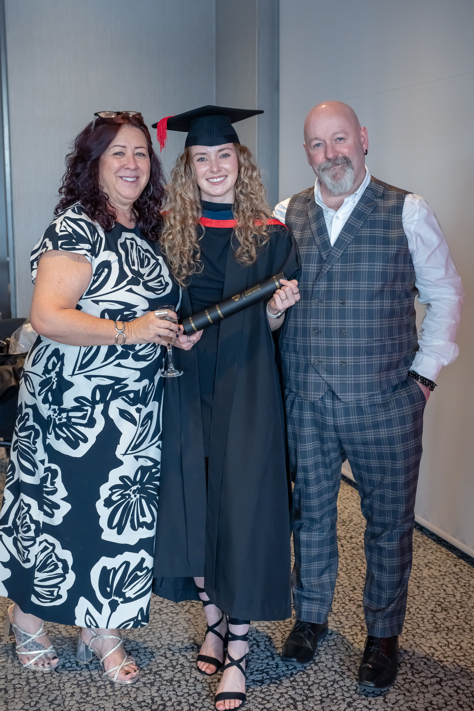 graduate smiles with parents. Graduate wears graduation cap and gown and holds a black scroll