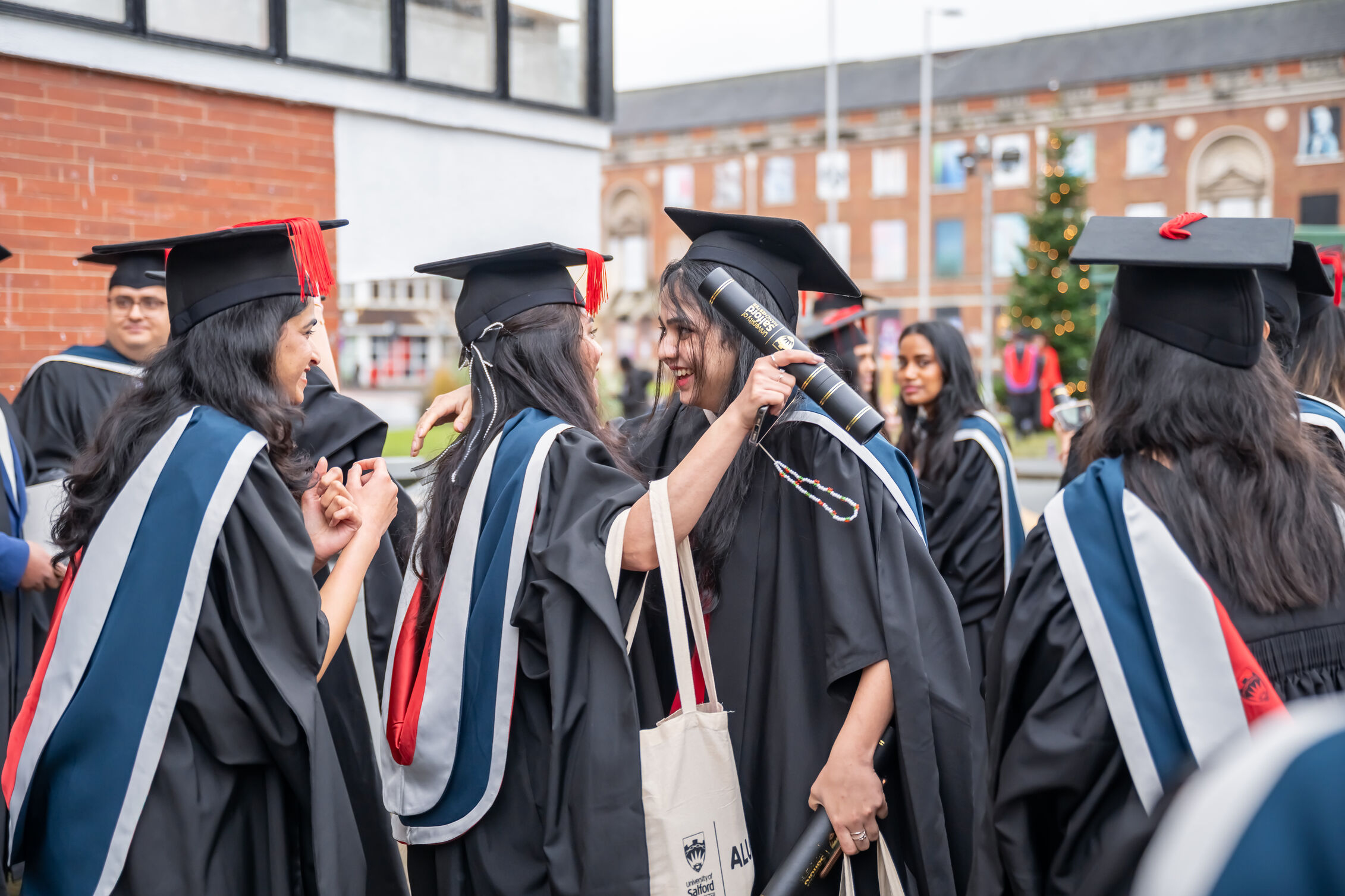 Graduates in black gowns and red, blue and grey hoods, chat and hug on Peel Park Campus. 