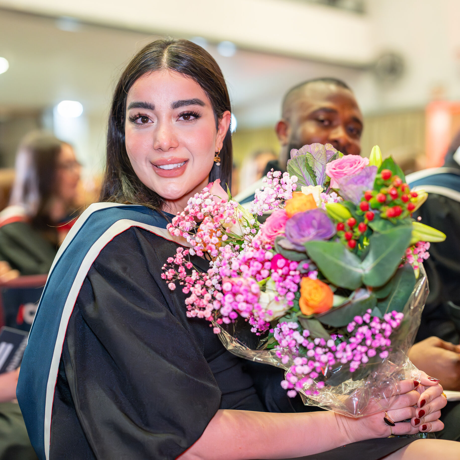 A graduate holds a bouquet of pink and orange flowers and smiles at the camera while sitting in the audience. She wears a black graduation gown and a blue and red hood.