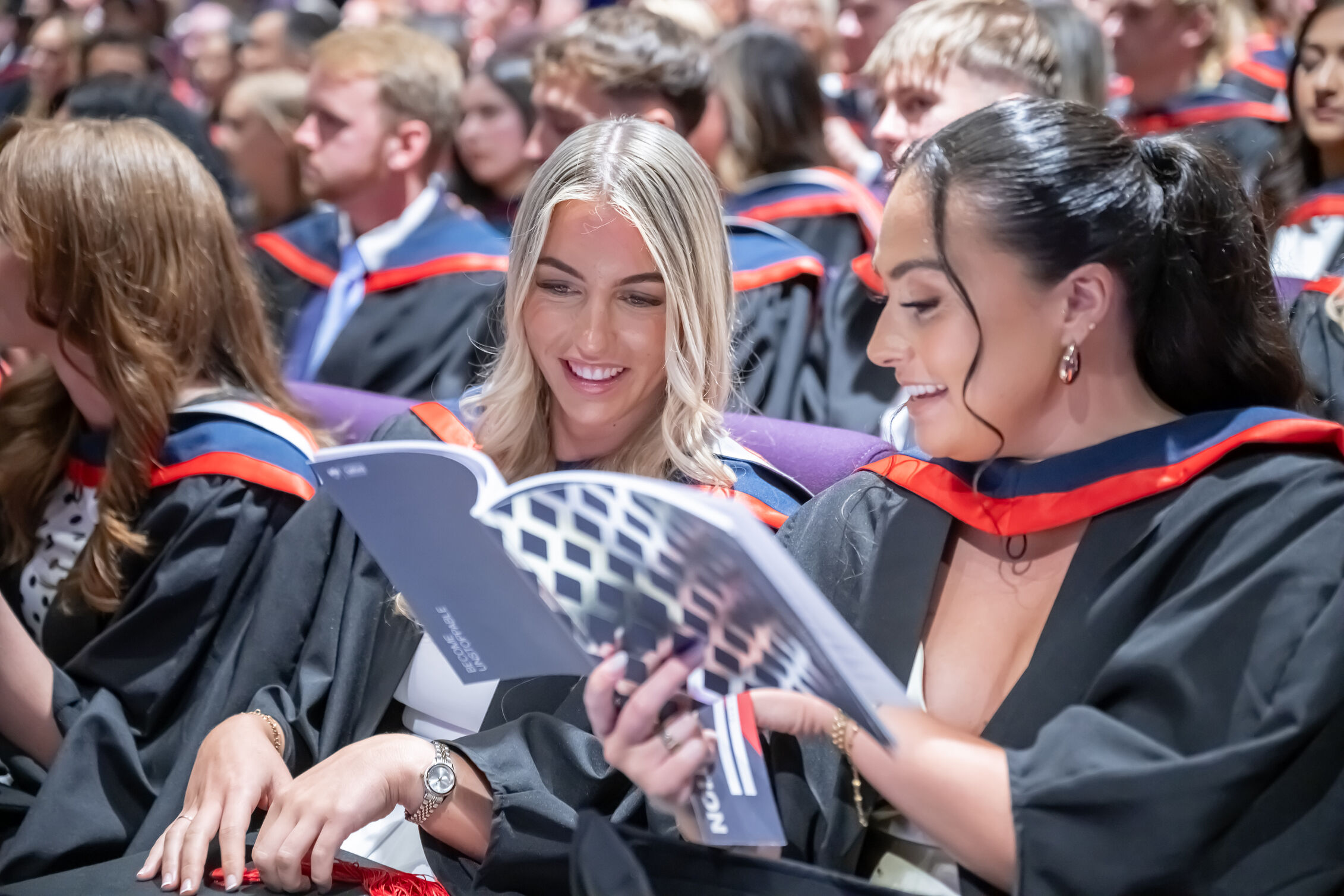 Two graduates read a Graduation brochure which is navy blue with a silver diamond pattern on.