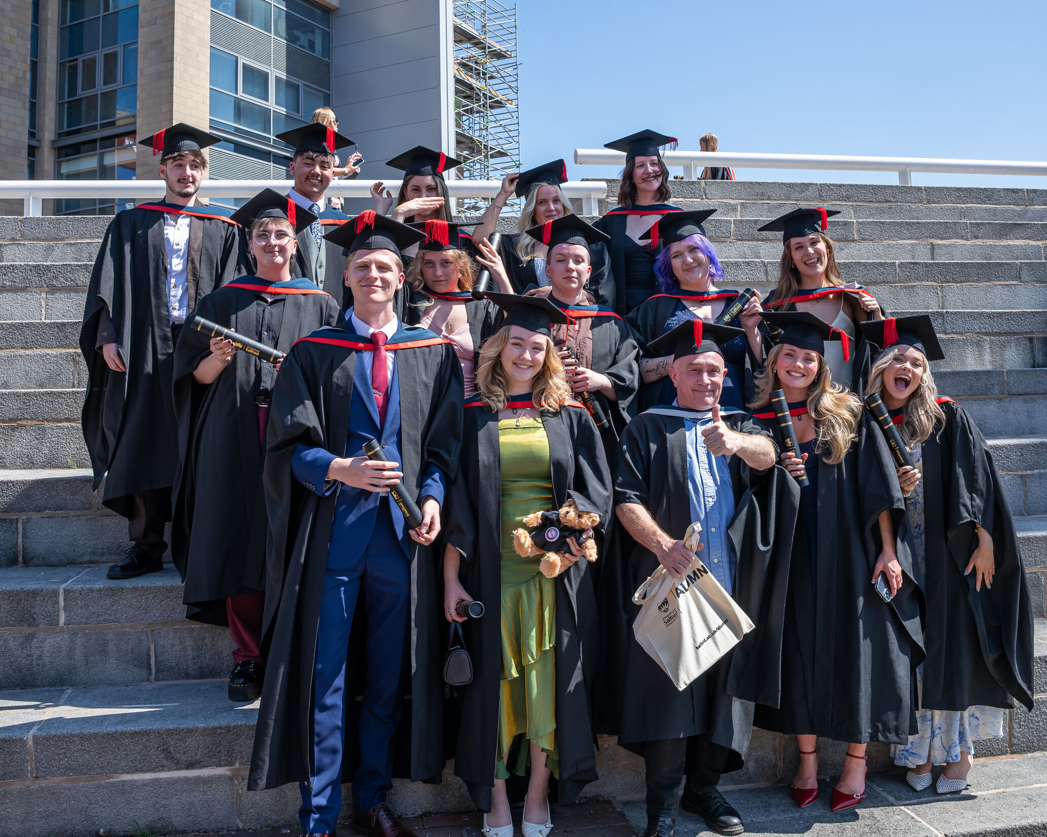 A group of graduates in black graduation gowns and black mortar board hats with red tassels smile outside on a sunny day. They hold various merchandise, including teddy bears with graduation caps on, black scrolls and alumni tote bags.