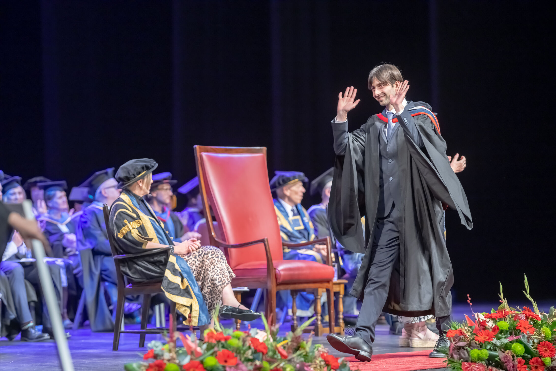 A graduate in a suit and black graduation gown raises his hand to wave ahead as they cross the stage. 