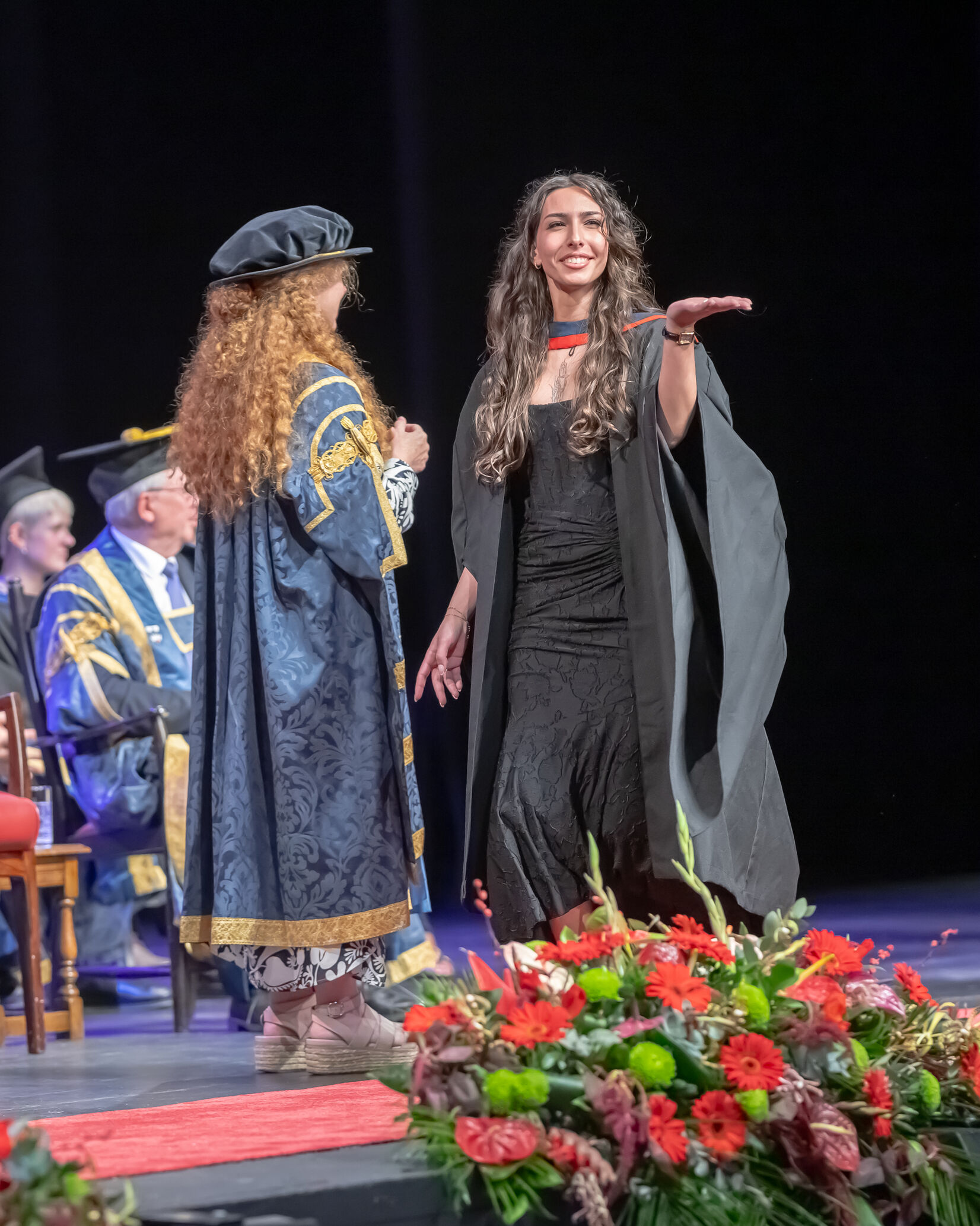 A graduate in a long black dress, black graduation robe and red and dark blue hood crosses the stage. They blow a kiss to the audience as they pass the Presiding officer, who wears a blue and gold gown and black bonnet.