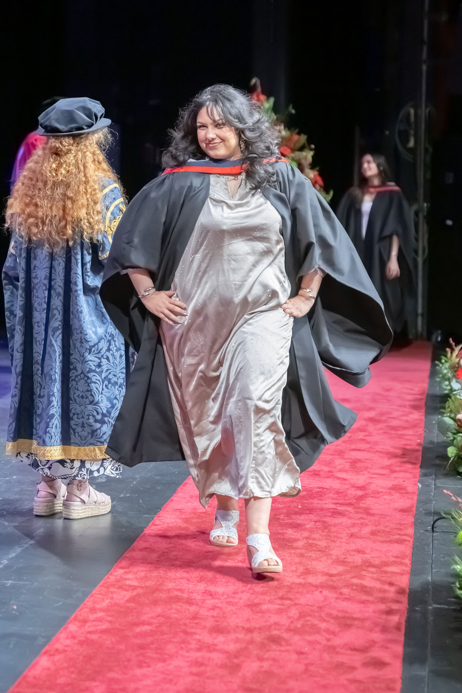 A graduate in a black gown and long silver dress, puts her hands on her hips and poses as she walks towards the camera on stage. The presiding officer can be seen behind her wearing a blue and gold gown.