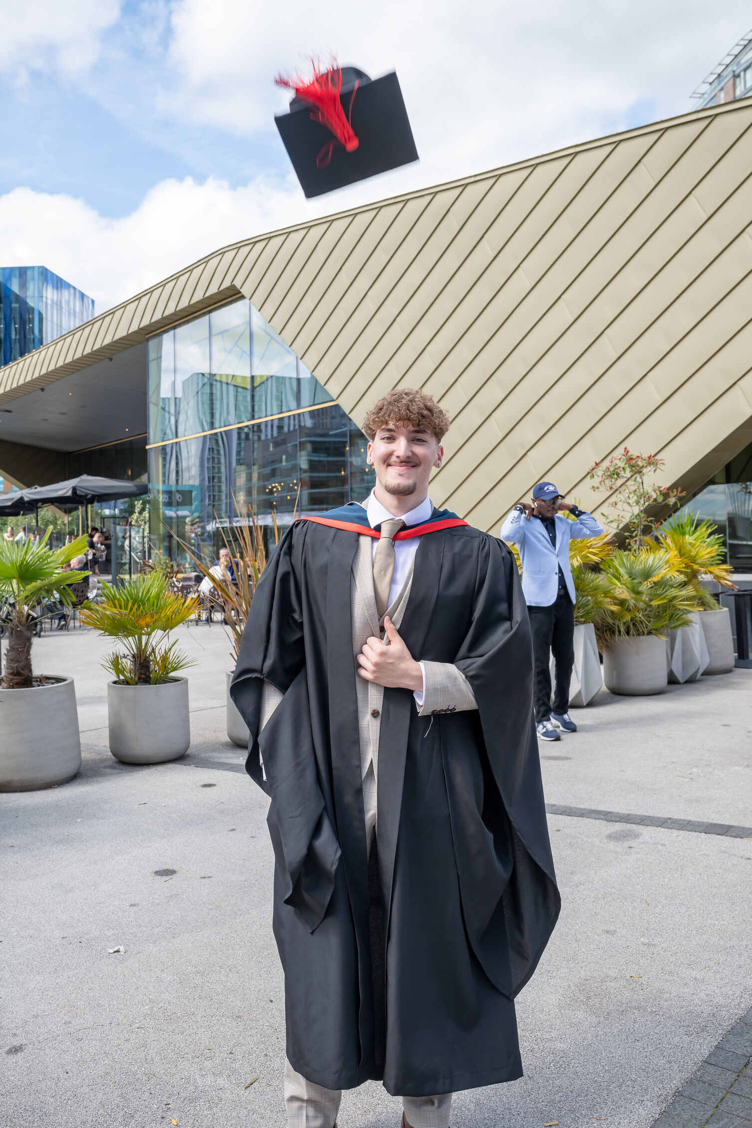A graduate wears a black graduation gown with a dark blue and red hood. They wear a  tan suit and tie under the gown. Their black mortar board hat with a red tassel hovers in the air above them after being thrown.