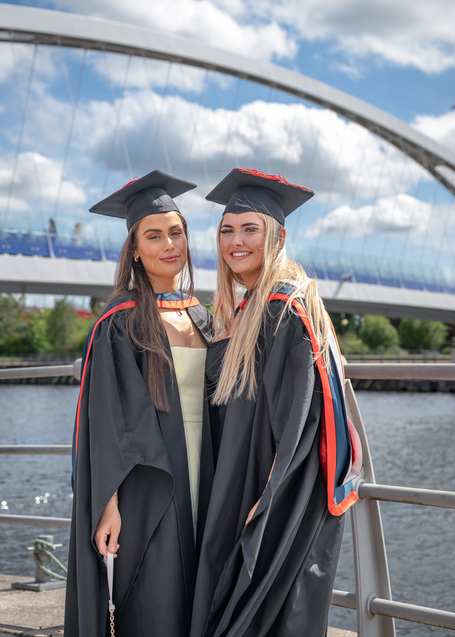 Two students in black graduation gowns, and dark blue hoods with blue trims smile at the camera. They stand in front of the Millennium Bridge at MediaCityUK. Both wear black mortar board hats with red tassels. The graduate on the left wears a yellow dress under her gown.