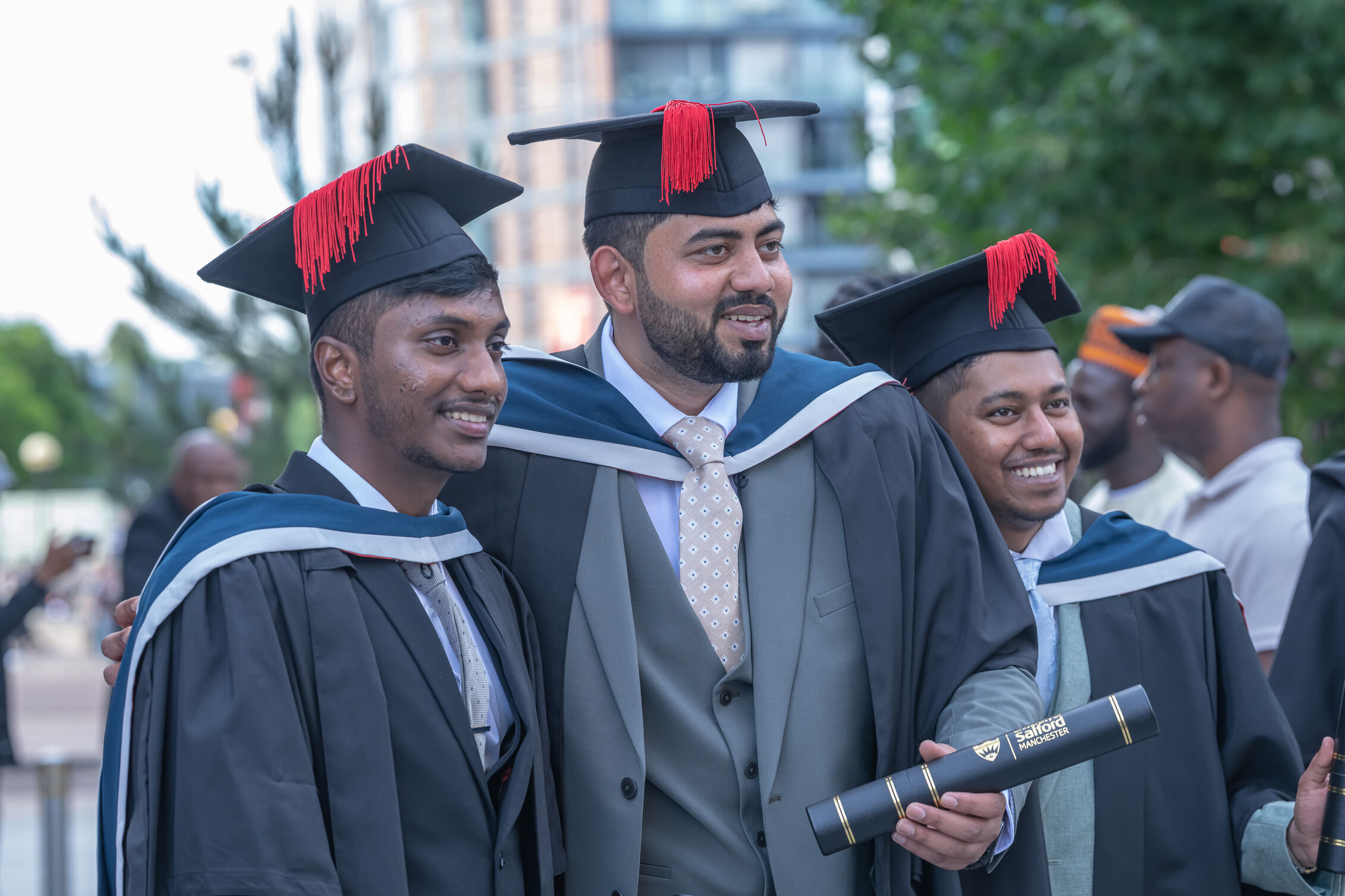 Three graduates in suits and black graduation gowns smile. All wear black mortar board hats with red tassels. The middle graduate holds a black scroll with a gold University of Salford logo on it. 