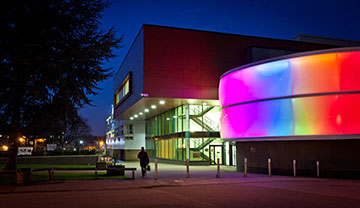 The Lady Hale building, a modern university building at night with a curved exterior wall illuminated by colorful lights, and a person walking nearby.