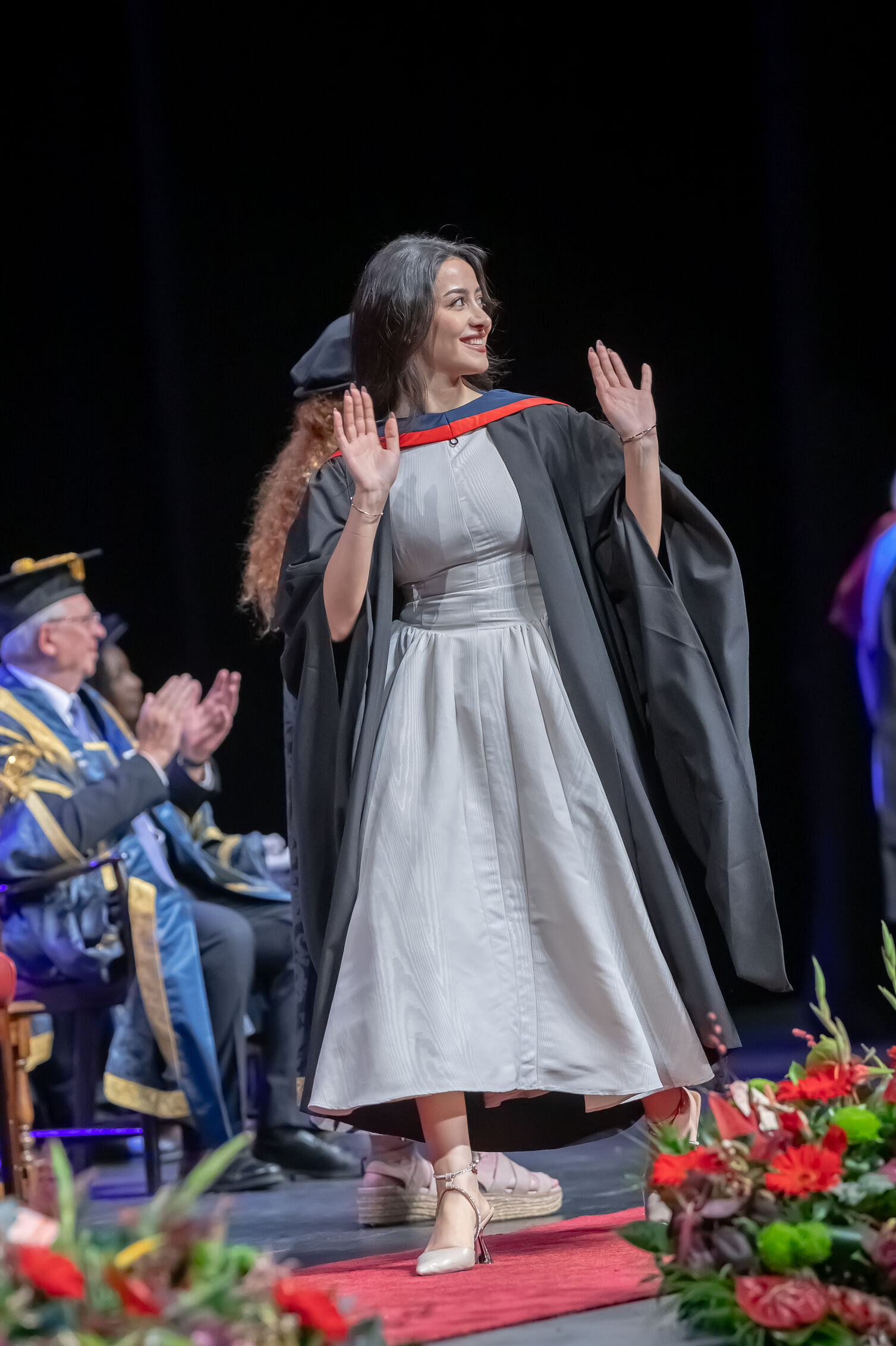 Graduate wearing a gown and red‑trimmed hood walks across a stage with hands raised, while seated officials applaud. Floral arrangements line the front of the stage.
