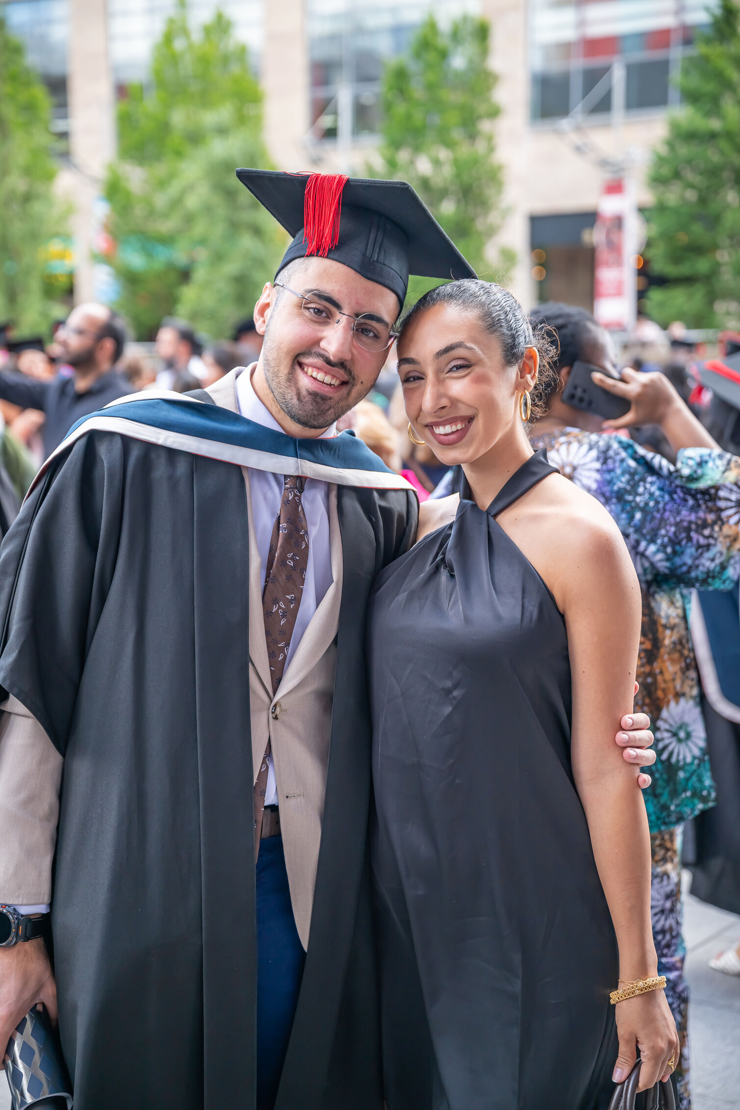 Two people pose together at an outdoor graduation event; one is wearing a cap, gown, and hood, and the other is dressed in a sleeveless black outfit. Trees, graduates, and guests are visible in the background.