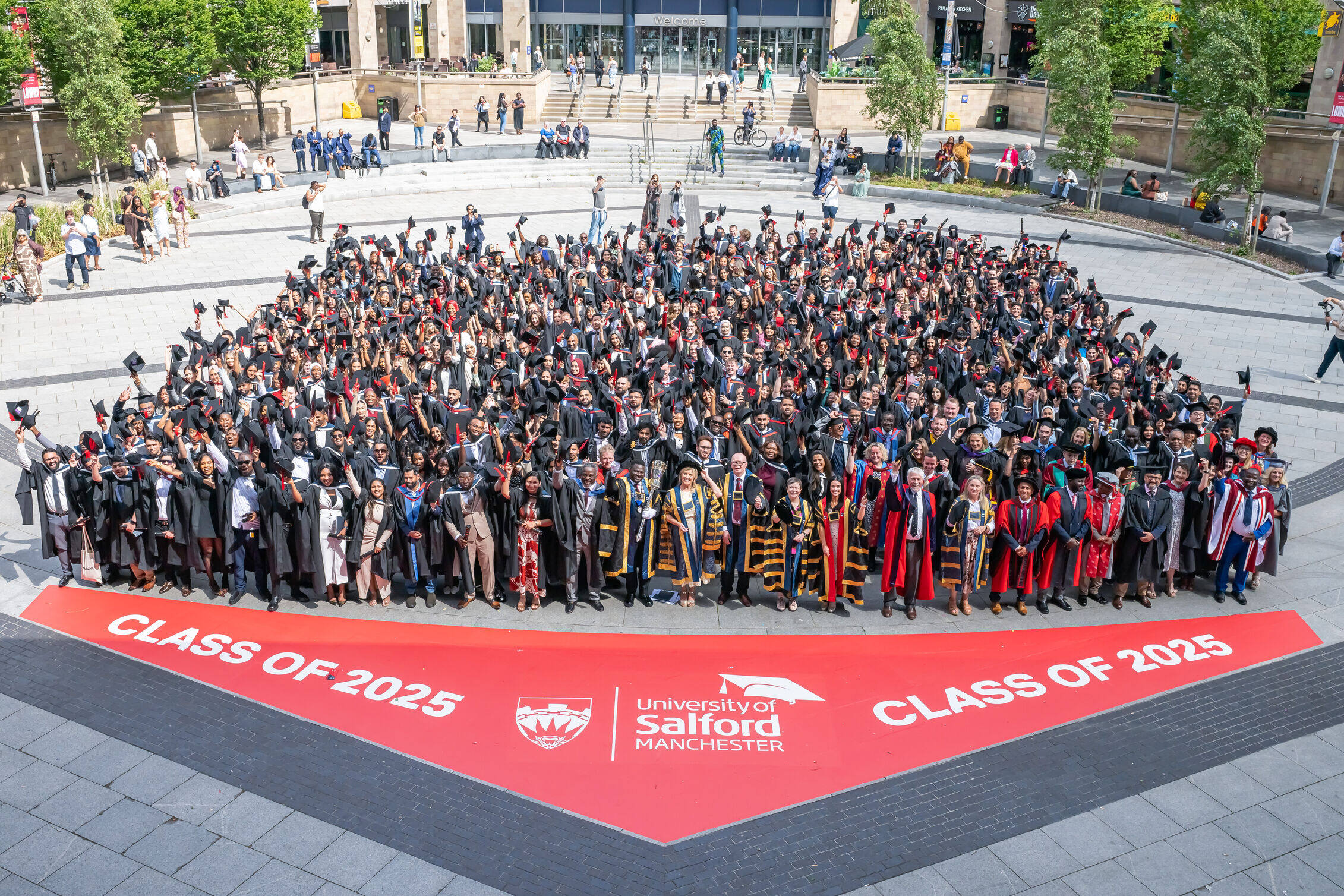 Large group of graduates in caps and gowns gathered in an outdoor square for a class photo. They are arranged in a wide triangular formation behind a red banner that reads ‘Class of 2025’ with University of Salford Manchester branding. Trees and university buildings surround the area.