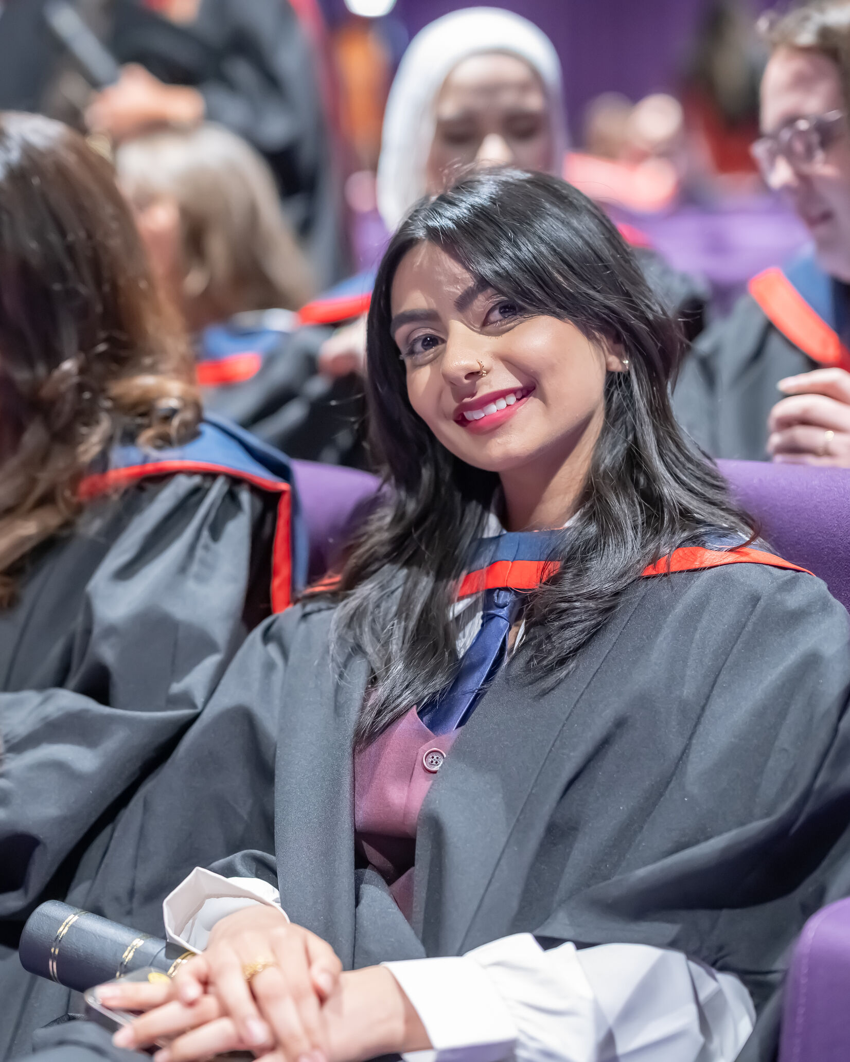 Graduate in a black gown with a red‑trimmed hood sits in a purple auditorium seat, holding a rolled certificate and smiling. Other graduates in academic dress are seated nearby.