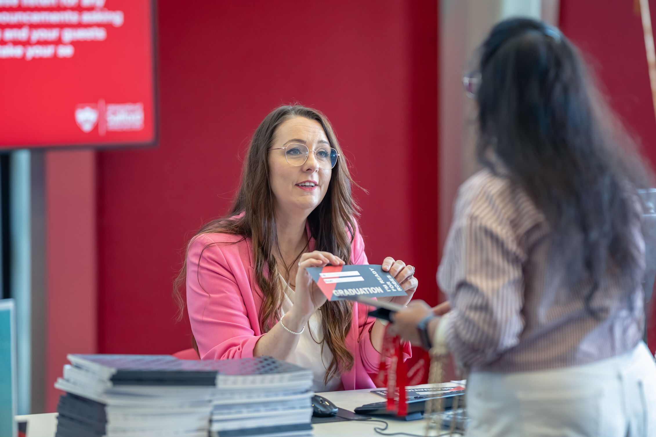 Staff member at a check-in desk hands a ticket that reads 'Graduation,' to a visitor across the counter. Brochures are stacked on the desk and a red display screen visible in the background.