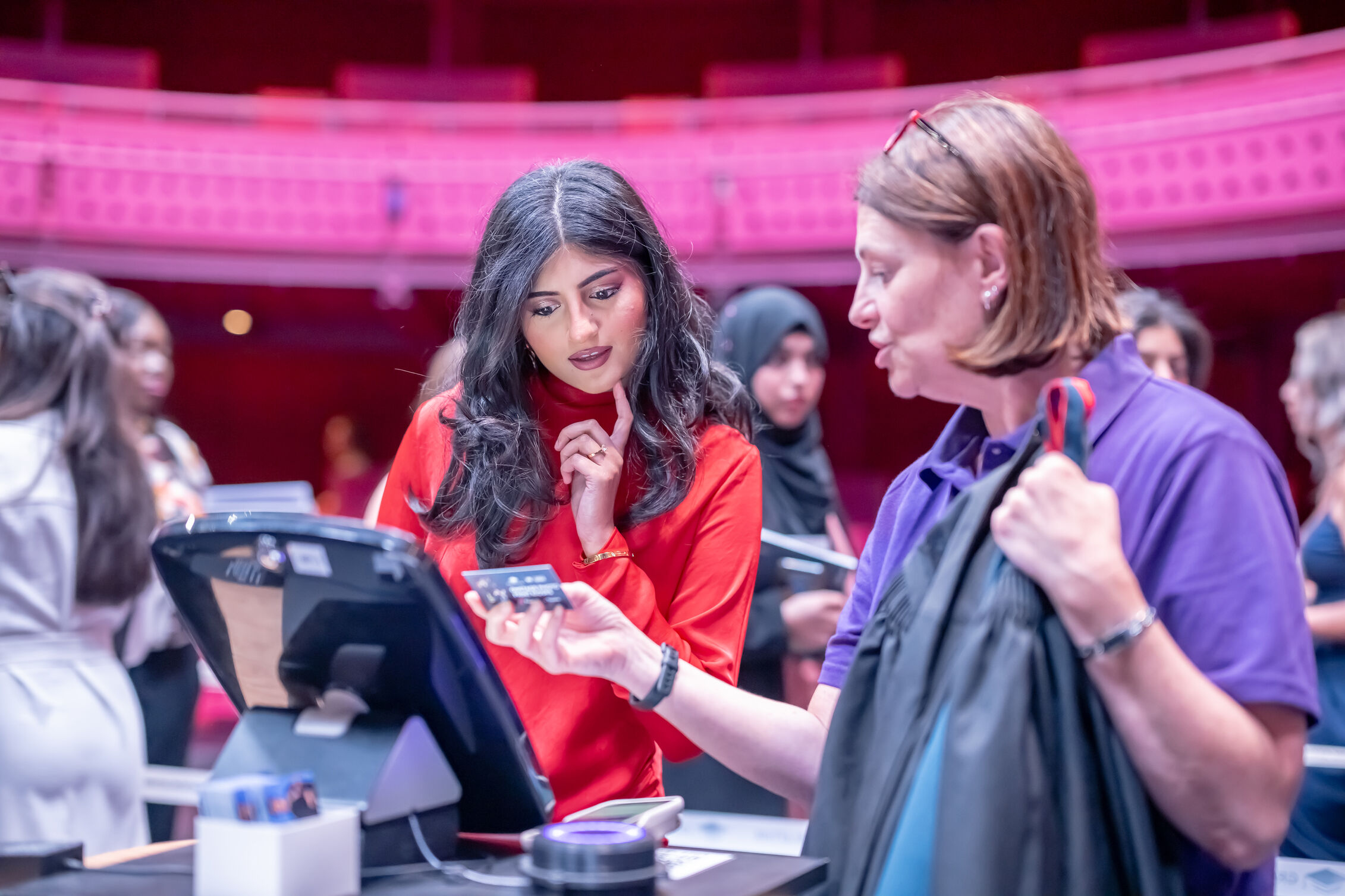 A staff member at a counter assists a visitor holding a card and a graduation gown. The staff member is using a touchscreen device, while people gather around in a brightly lit indoor venue with pink and purple lighting.