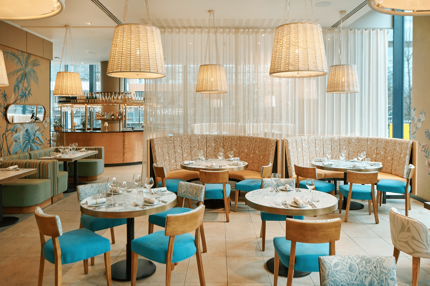 A bright, modern restaurant interior featuring round tables set for dining, curved upholstered booths, wicker pendant lights, and floor‑to‑ceiling sheer curtains. The space has warm wood accents and a contemporary, inviting atmosphere.