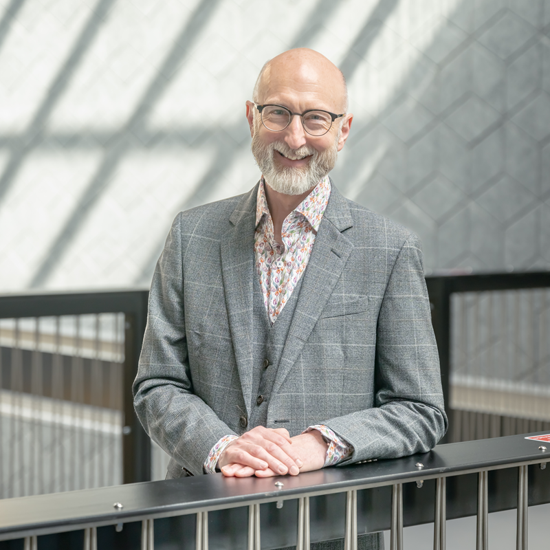 Man with glasses and beard, wearing grey suit and patterned shirt, smiling for photo on indoor balcony