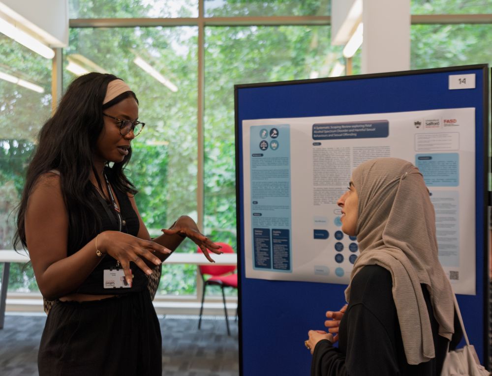 Two women speaking in front of a presentation board that is showing research