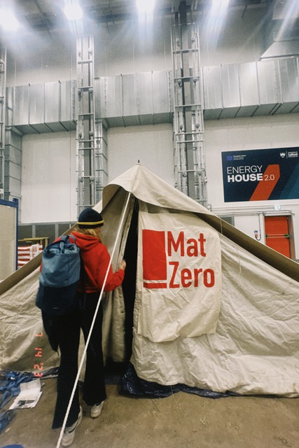 There is someone in the centre of the image lifting a pole up. They are putting up a tent that has a logo saying 'MAT' on the side of it. Th person has a red coat and a blue bag on their back. They are doing this within the energy house building, which is a big building with white walls and there is an energy house sign in the background.
