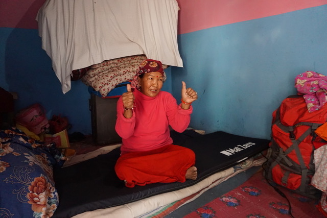 A Nepali lady sat in her house, in a pink top and pink skirt, on top of a black mat. She has her thumbs up.