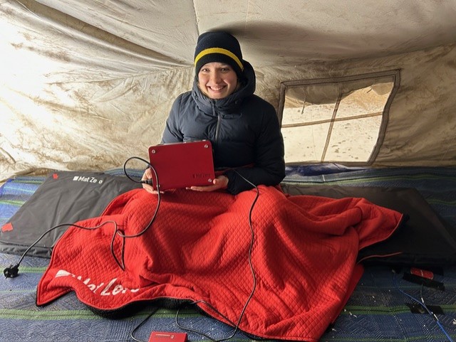 A lady with a coat and a knitted hat on, smiling with a red blanket over her. She is in a tent and has a laptop on her knee.