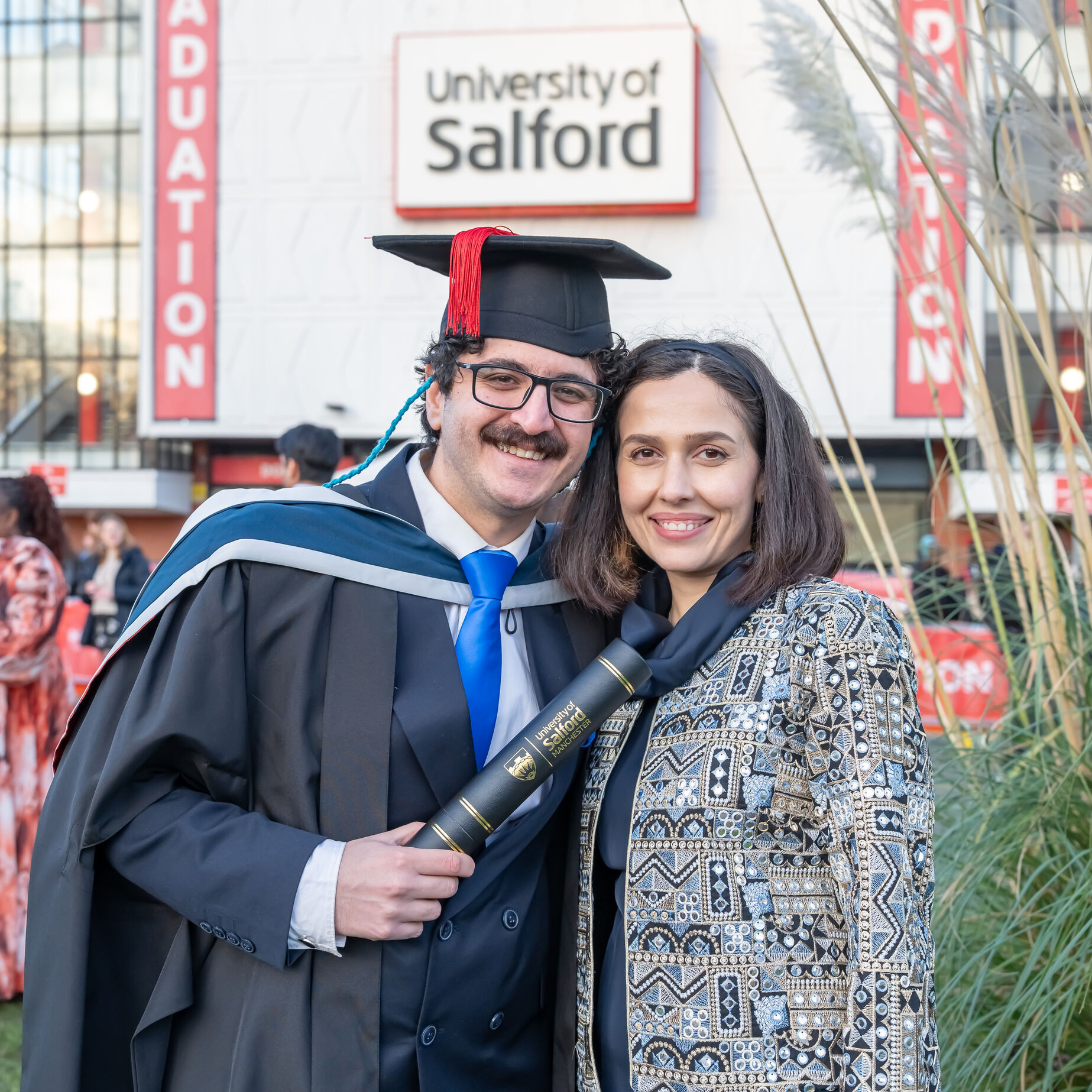 Graduate in academic dress holding a degree certificate alongside another person in front of Maxwell Building, University of Salford