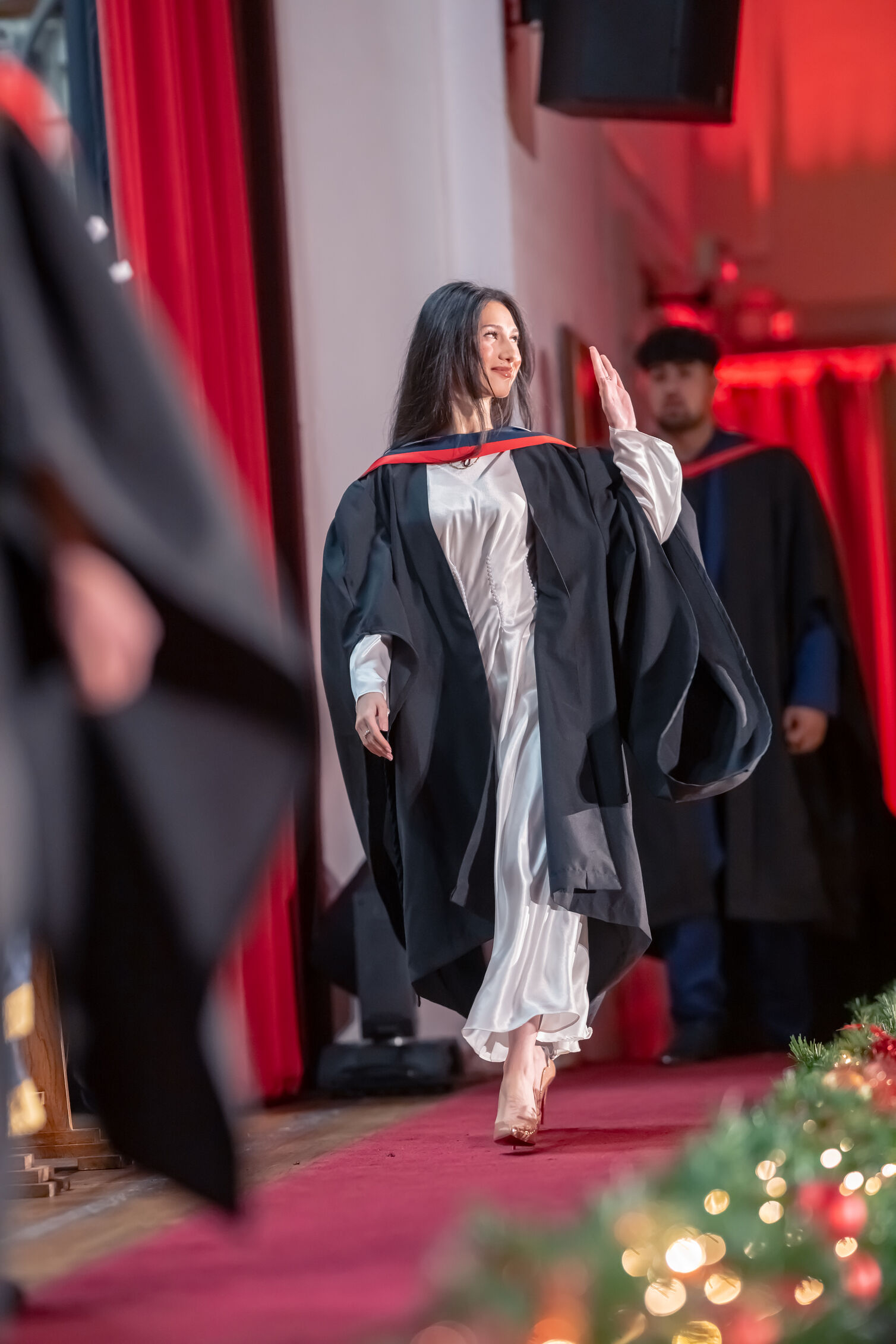 Graduate in academic dress walks across a stage, raises a hand in acknowledgement during a graduation ceremony