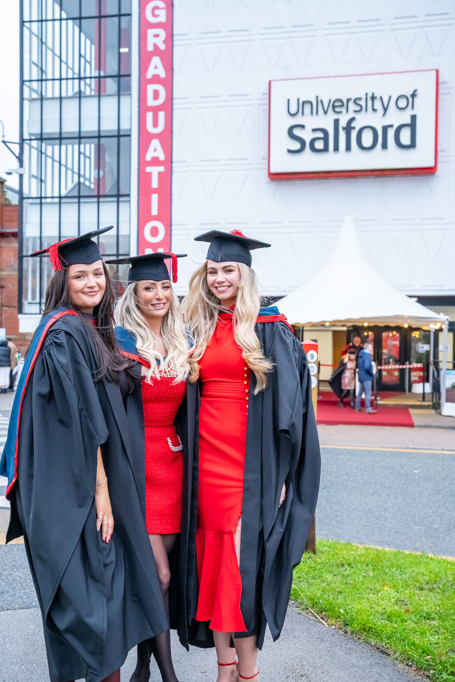 Three graduates wearing academic gowns and mortarboards pose for a photograph outside a graduation venue. Two graduates wear bright red dresses beneath their gowns, while the third wears a dark outfit. Behind them, the building displays large signs reading “GRADUATION” and “University of Salford,” with the entrance tent and arriving guests visible in the background.