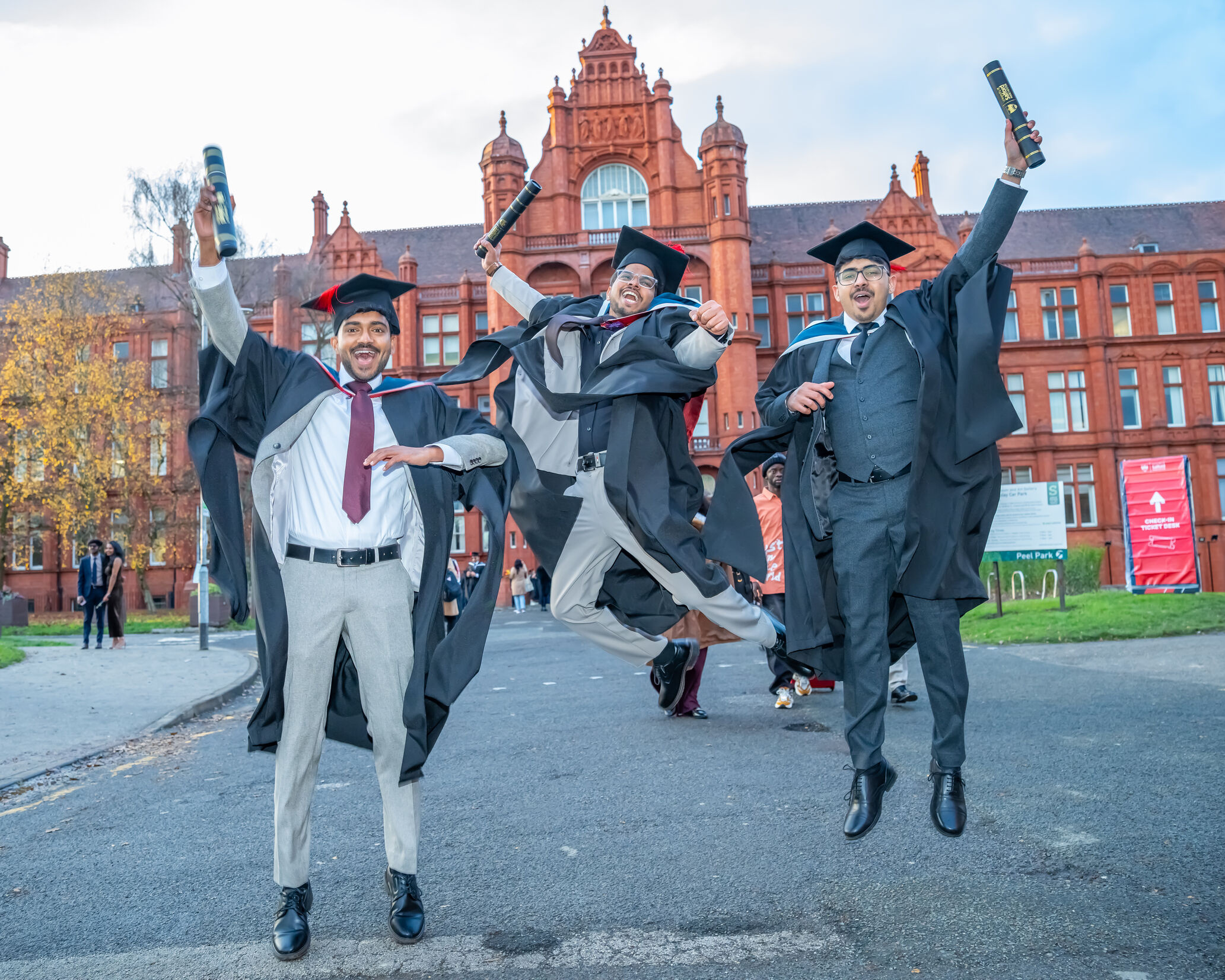 Three graduates in academic dress jump into the air while holding diploma tubes in front of Peel Building, University of Salford