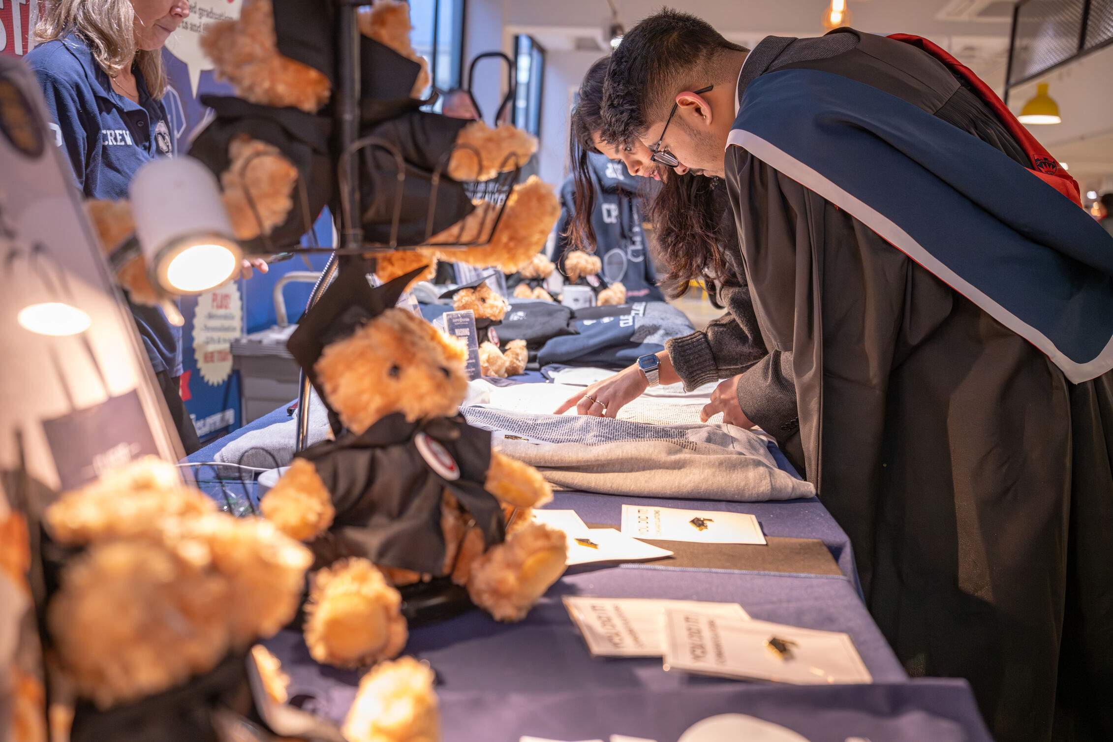 Graduates in academic regalia browsing merchandise including souvenir teddy bears 