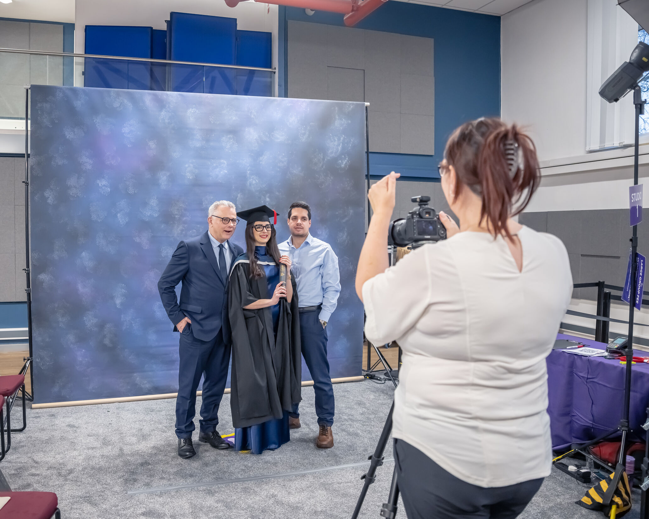 A graduate wearing academic robes and a mortarboard stands between two guests while holding a diploma tube and posing for a photograph in a graduation photo studio. In the foreground, a photographer operates a camera on a tripod. The group stands in front of a blue mottled backdrop inside a venue with studio lighting, seating, and event equipment visible around the photography area.