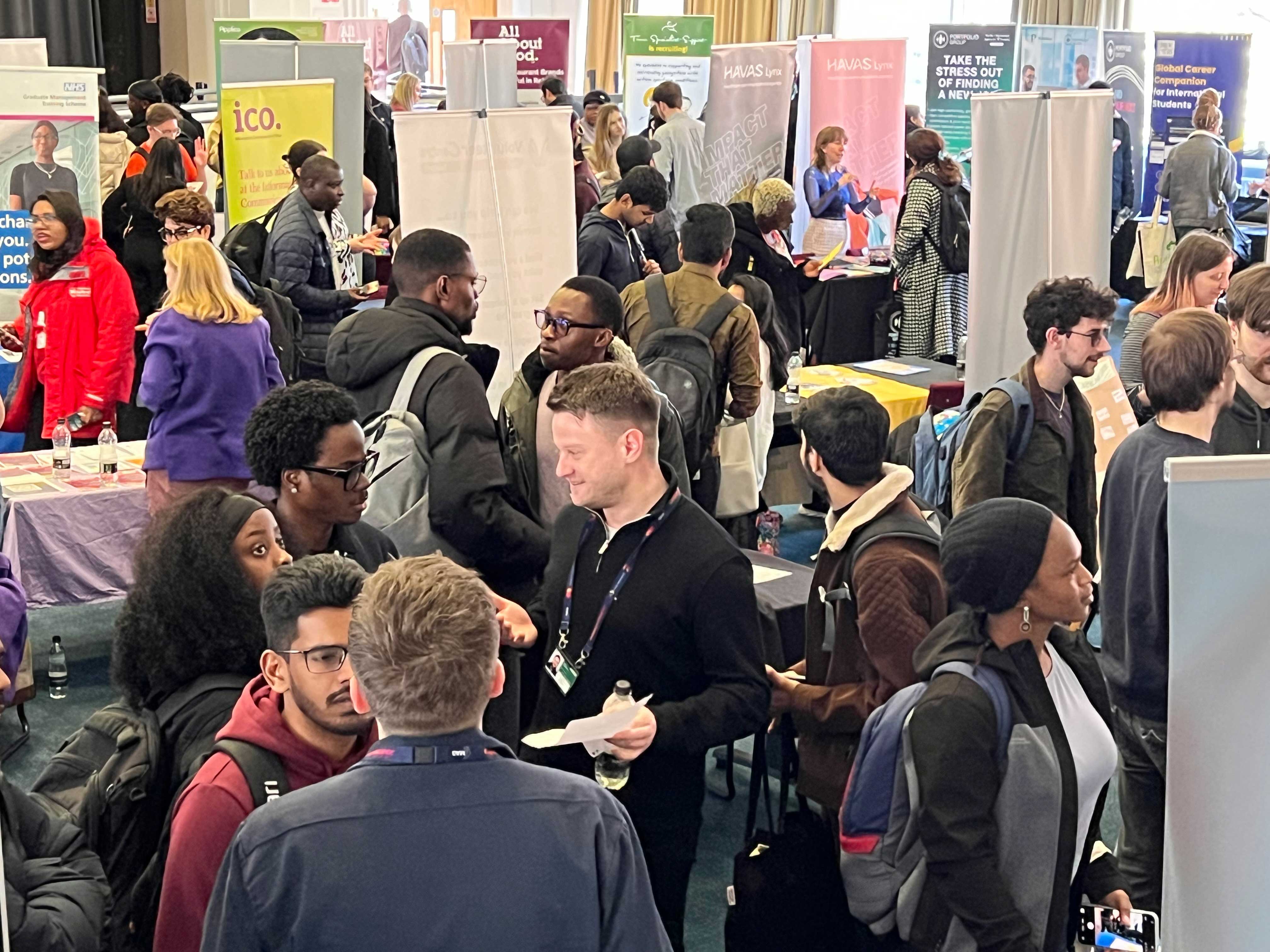 Crowded careers and recruitment fair with attendees speaking to representatives at exhibitor stands and information booths in a large indoor venue
