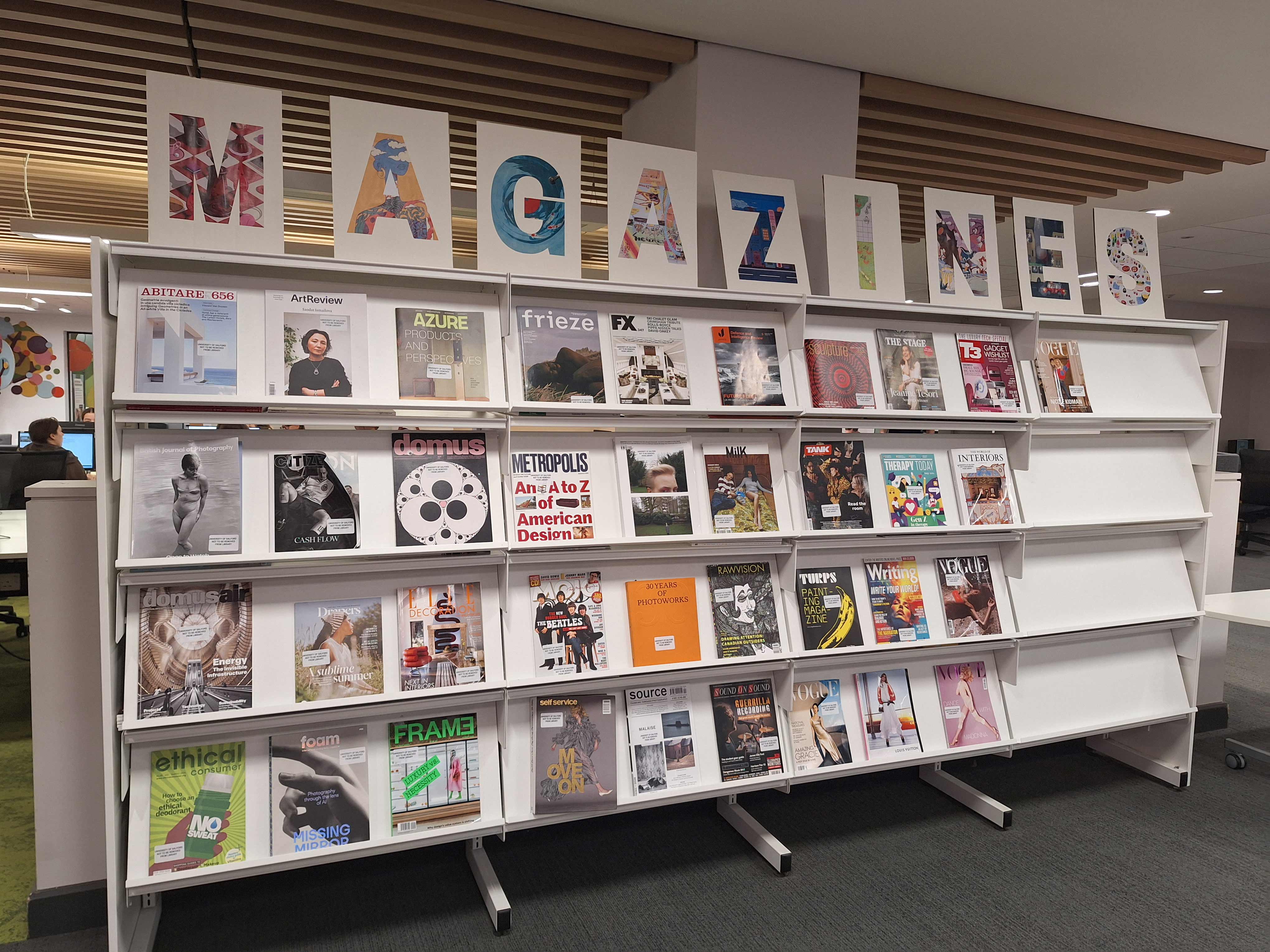Library-style magazine display in an office workspace, showing rows of art, design and lifestyle magazines on white shelving. Large decorative letters spelling “MAGAZINES” are mounted above the display